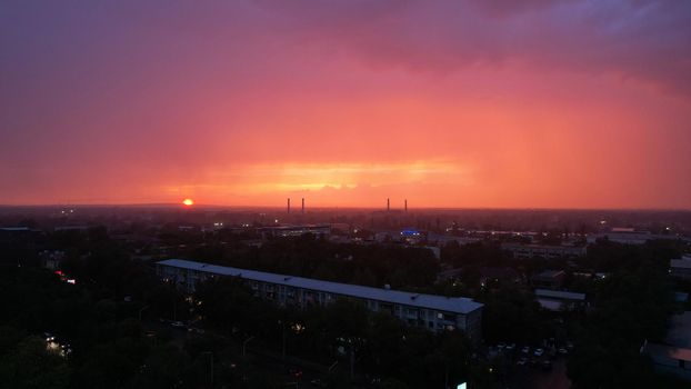 Epic sunset with clouds over the city of Almaty. The gradient of clouds is from dark blue to purple-orange. Green tall trees, houses, a road with cars. The lights are on. It's raining heavily