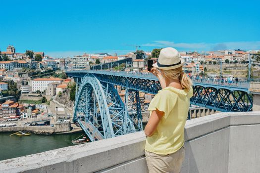 Blonde girl in a hat girl makes selfie on smartphone near Dom Luis Bridge in Porto, Portugal