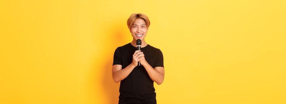 Portrait of charismatic smiling asian man holding microphone and singing karaoke, performing song, standing yellow background.