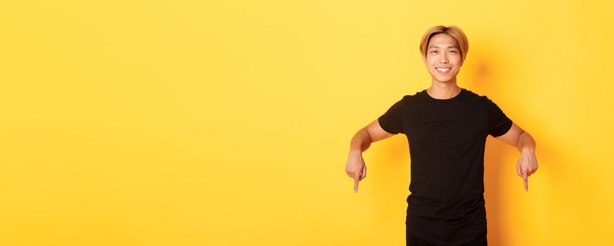 Portrait of smiling attractive korean guy with blond hair, wearing black outfit, pointing fingers down, showing way, standing yellow background.