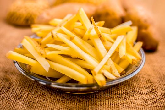 Raw cut french fries in a transparent glass plate along with raw potato with it on jute bag's surface.