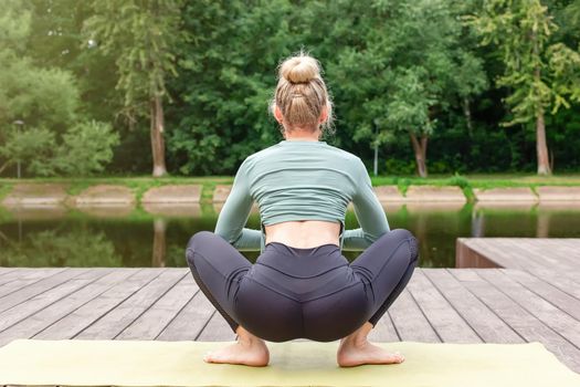 A slender woman in a gray top and leggings, sitting on a wooden platform by a pond in the park in summer, does yoga on green mat . Back view. Copy space