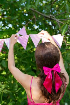 The girl hangs triangular flags of pink color against the backdrop of a green garden. A girl in a pink dress and a bow on her hair. Festive decor - celebration concept