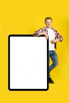 Pointing finger handsome caucasian man leaned on giant, huge digital tablet with blank white screen, wearing casual outfit isolated on yellow background. Free space mock up.