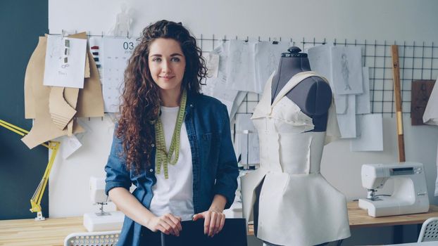 Waist-up portrait of beautiful young woman tailor standing in workplace near clothed mannequin and looking at camera. Women's garments, sketches and sewing machines are visible.