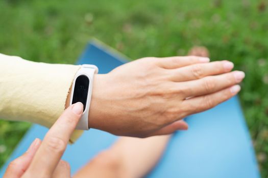 Sports girl doing stretching in nature outdoors, the girl looks at her fitness bracelet and checks her breathing during exercise. A woman is doing yoga in the park, sitting on the grass on a yoga mat