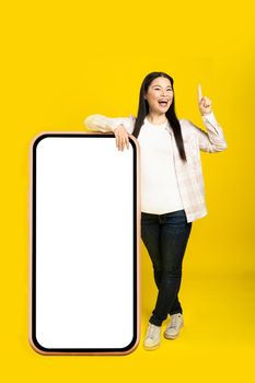 Middle age asian woman excited pointing finger up leaned on giant, huge smartphone with blank white screen, wearing casual outfit isolated on yellow background. Free space mock up.