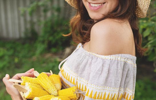 The girl is smiling in a straw hat and holding a plate of boiled corn in her hands. The concept of outdoor recreation, barbecue. Noise effect