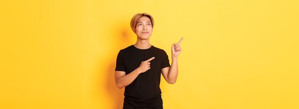 Thoughtful and pleased handsome asian man smiling and pointing fingers upper left corner, standing over yellow background.