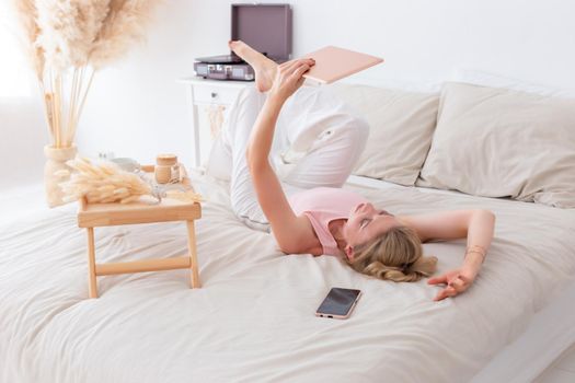 A slender woman, 30-40 years old, with blond hair, in a pink top and white pants, lies on her back in the morning on the white bed, legs up, holds a pink digital tablet in her hands.