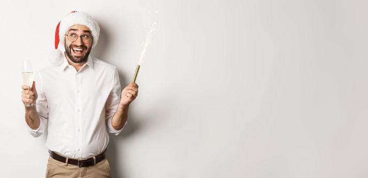 Winter holidays and celebration. Happy businessman enjoying New Year party, wearing Santa hat and drinking champagne, smiling amused, white background.