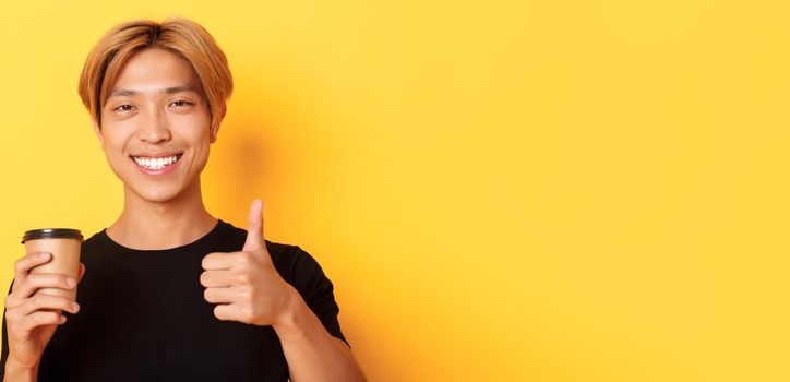 Close-up of satisfied asian handsome guy recommend cafe, holding cup of coffee and showing thumbs-up in approval, smiling pleased over yellow background.