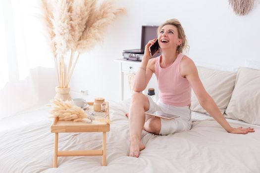 Happy woman with blonde hair, in a pink top and white pants, sitting on the bed in the morning, in a bright living room interior, talking on the smartphone and holding a digital tablet on her knees. Copy space