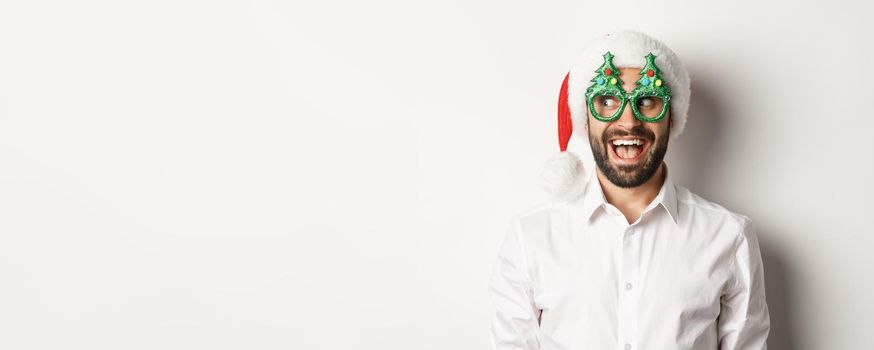 Close-up of funny man looking left with surprised face, wearing christmas party glasses and santa hat, celebrating New Year, white background.