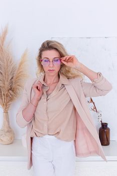 Portrait of stylish woman in glasses, with blond hair stands in a light pink jacket in the interior of a white room, look at down . Copy space. Vertical