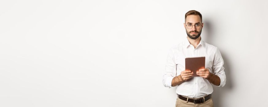 Serious employer working with digital tablet, reading in glasses, standing over white background.