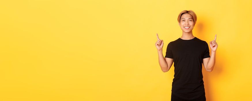 Portrait of handsome smiling asian guy in black t-shirt pointing fingers up and looking satisfied, yellow background.