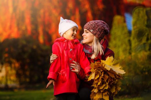 Mother drinking tea with kid daughter 4-5 year old sitting in sun light in park. Wearing autumn knitted clothes. Looking at each other. Motherhood. Childhood.