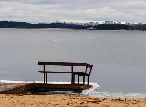 Two wooden benches on small pier on sandy lake shore in winter. Wooden bench on the pier near the shore. A pier on the winter seashore.