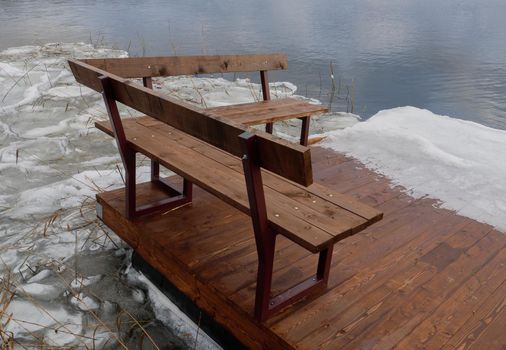 A small wooden pier on the ice of the freezing sea. Bench on the winter shore of the lake. A pier on the winter seashore.