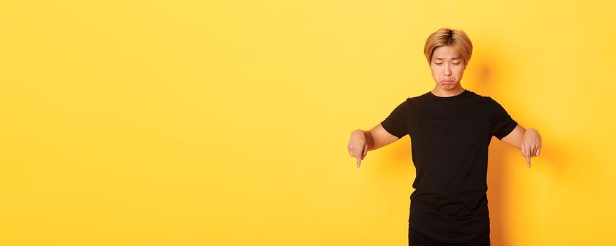 Portrait of gloomy and upset asian man sulking with regret, pointing and looking down at banner, yellow background.