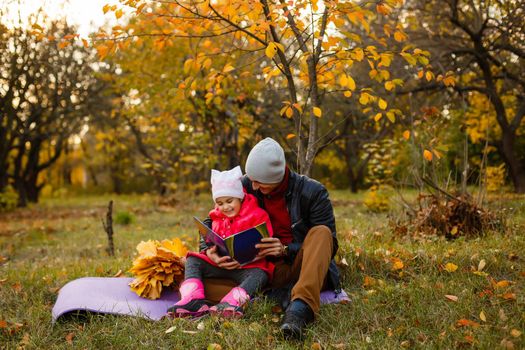Happy father and daughter sitting and reading a book in autumn park on yellow leaves