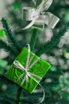 A green gift box tied with a white ribbon hangs from a green holiday tree.