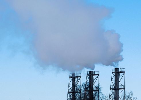 Three factory chimneys against a blue sky with smoke.