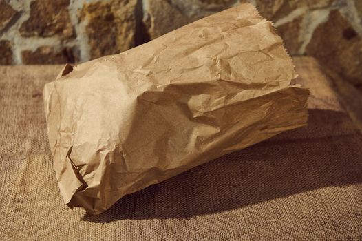 Close-up of an eco-friendly recyclable craft paper bag with healthy wholegrain wheat bread inside, on a table with a sackcloth tablecloth. Copy advertising space. Selective focus