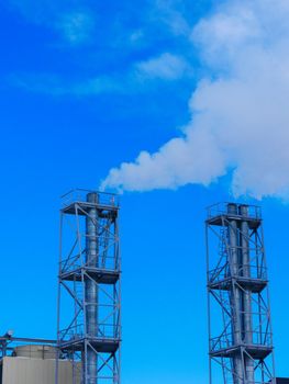 Two chimneys of a heating plant against the background of a bright blue sky and smoke coming out of them.