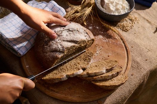 View from above of hands cutting a homemade whole grain round loaf of healthy sourdough rye bread on a wooden board. Artisanal bakery