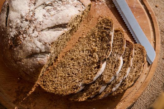 Closeup of a homemade delicious whole grain fresh baked sourdough rye bread, with crumbs while being sliced on a wooden cutting board. View from above. Still life with food. Concept of bakery