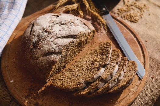 View from above of slices of freshly baked whole grain sourdough bread on a wooden board on a table covered with a burlap tablecloth, wheat ears and scattered cereal grains on a blurred background
