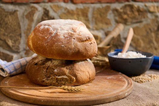 Still life with fresh baked whole grain, multigrain sourdough loaves of bread on wooden board, a bowl with white flour on table covered with burlap tablecloth against stone wall background, close up