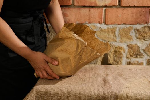 Selective focus. Eco bag with fresh whole grain healthy baked pastry, rich in nutrient fibers, in the hands of a baker confectioner wearing black chef's apron standing at table with burlap tablecloth