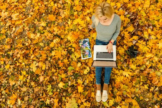 Cute woman with laptop in the autumn park. Beauty nature scene with colorful foliage background, yellow trees and leaves at fall season. Autumn outdoor lifestyle. Happy smiling woman on fall leaves