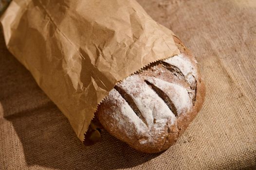 Still life with an artisanal whole grain, healthy rye sourdough bread, in a recyclable eco paper bag on the rustic table with sackcloth tablecloth against a brick and stone wall background. Top view