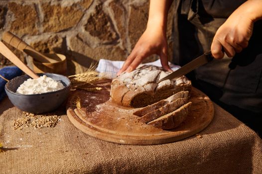 Selective focus. Baker cuts a loaf of multigrain sourdough rye bread on a wooden board. White flour and wheat spikelets on the table, covered with burlap tablecloth. Artisanal rustic bakery. Closeup