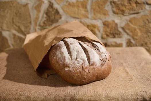 Still life with an artisanal whole grain, healthy wheat homemade sourdough bread, in a recyclable craft paper bag on the rustic table with burlap tablecloth against a brick and stone wall background