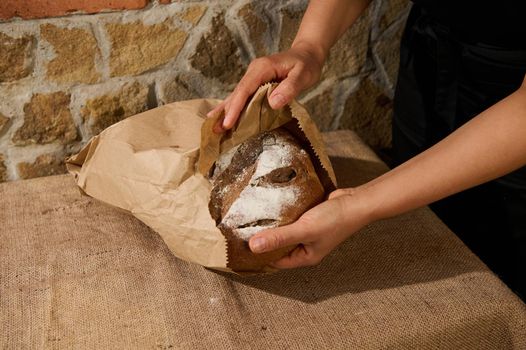 Details: Hands of baker confectioner putting fresh whole grain wheat homemade delicious bread, full of nutrient fibers and vitamins, into an eco recyclable paper bag. Bakery and healthy eating concept
