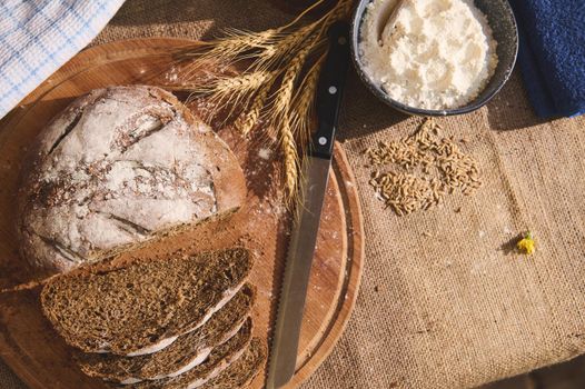 Still life with delicious healthy fresh baked homemade sourdough bread, sliced on wooden board, a kitchen knife wheat spikelets and white flour in blue ceramic bowl on the burlap tablecloth. Top view