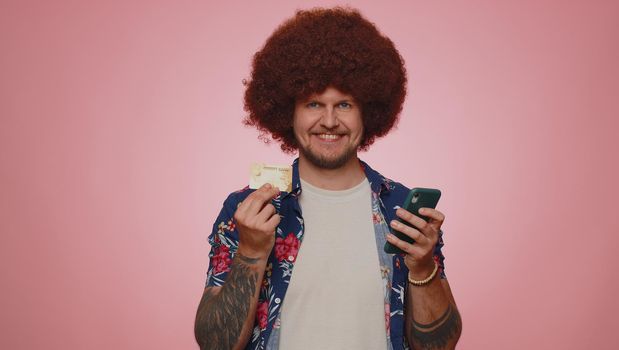 Sincere adult man customer using credit bank card and smartphone while transferring money, purchases online shopping, payment. Finance and internet. Young guy isolated alone on pink wall background