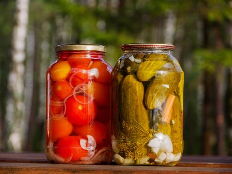 Two glass jars with pickled cucumbers and tomatoes outdoors.