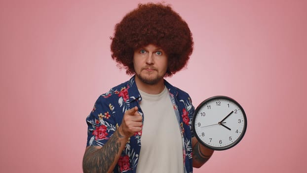 It is your time. Portrait of bearded young man in shirt showing time on clock watch, ok, thumb up, approve, pointing finger at camera. Adult guy indoors studio shot isolated alone on pink background