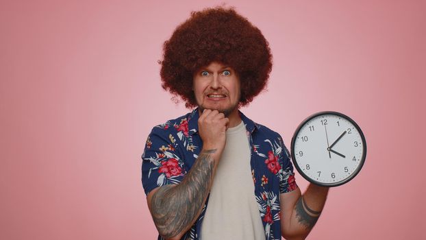 Confused young man with anxiety checking time on clock, running late to work, being in delay, deadline on pink studio background. Serious guy looking at hour and minutes, worrying to be punctual