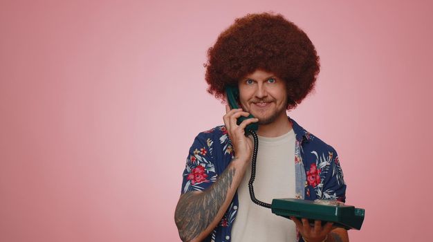 Hey you, call me back. Cheerful man secretary in blue shirt talking on wired vintage telephone of 80s, says hey you call me back. Young adult guy posing isolated on pink studio wall background