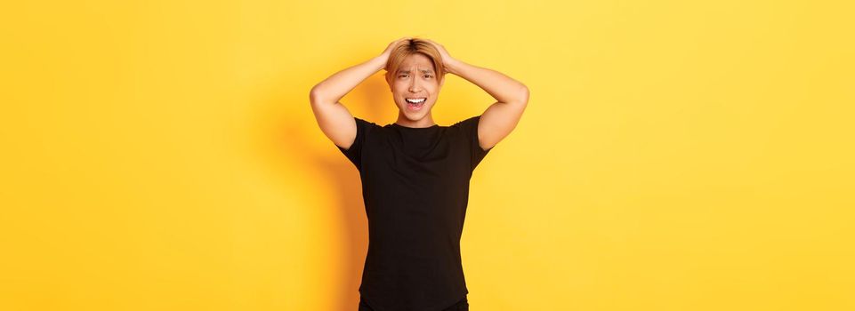 Portrait of young asian guy panicking, looking anxious and upset, holding hands on head and grimacing, standing over yellow background.