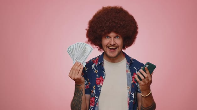 Happy tourist man looking smartphone display sincerely rejoicing win, receiving money dollar cash banknotes, success lottery luck. Young bearded guy boy isolated alone on blue studio background