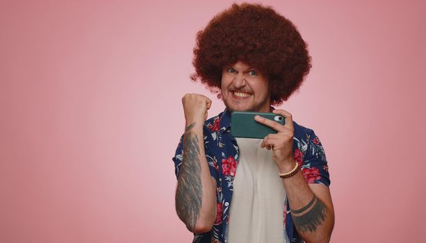 Worried funny addicted man in shirt enthusiastically playing racing video games on mobile phone. Young adult guy using smartphone gadget app with drive simulator isolated on pink studio background