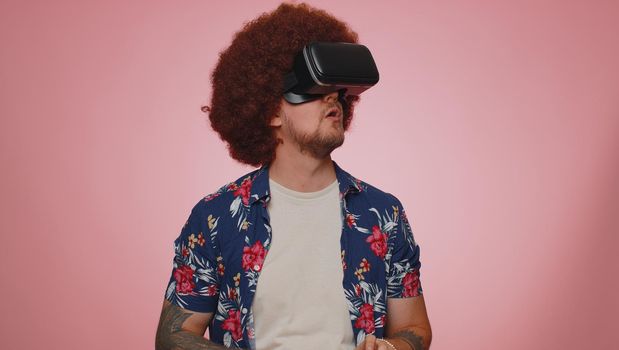 Exited stylish man using virtual reality futuristic technology VR app headset helmet to play simulation 3D 360 video game, drawing. Young adult guy on pink studio background. People sincere emotions